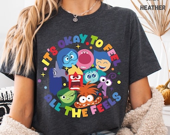 Könnte beinhalten: Dunkelgraues Heather-T-Shirt mit einem farbenfrohen Grafikdesign. Das Design zeigt Cartoon-Charaktere, die verschiedene Emotionen darstellen, mit dem Text "IT'S OKAY TO FEEL ALL THE FEELS" in einem Kreis.