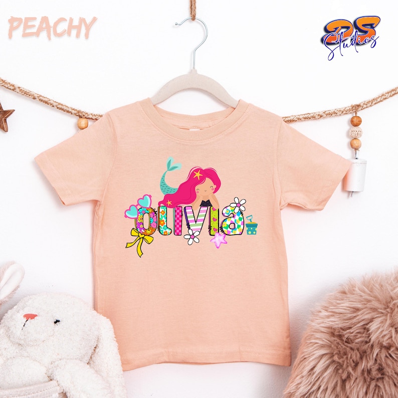 Puede incluir: Una camiseta de manga corta color melocot&oacute;n con el gr&aacute;fico de una sirena de pelo largo y rosa. El nombre OLIVIA est&aacute; impreso en letras grandes, coloridas y estampadas, decoradas con peque&ntilde;as flores, corazones y un lazo amarillo.