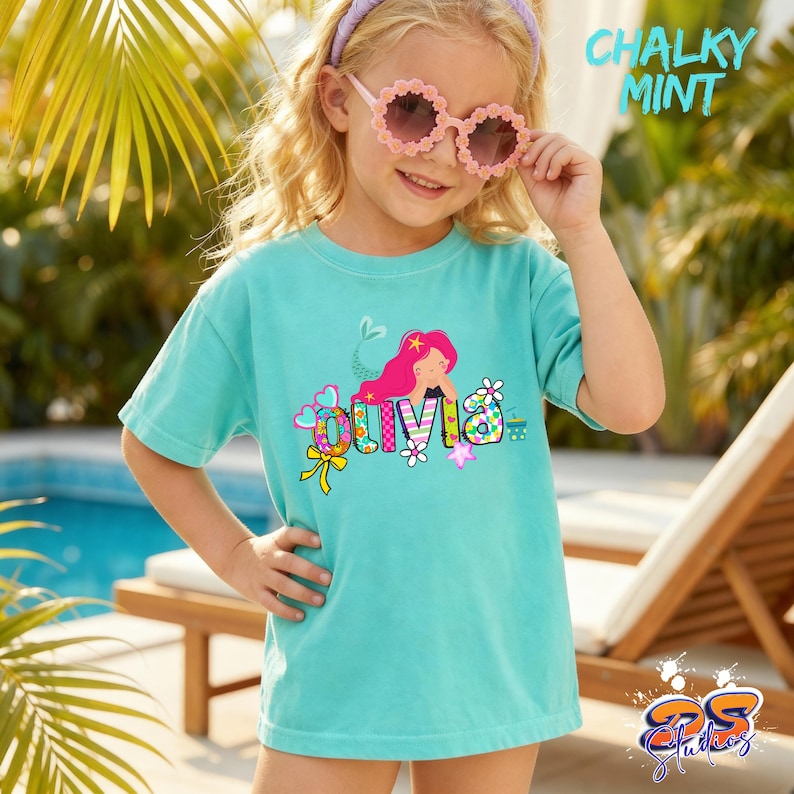 Puede incluir: Una camiseta de manga corta color menta tiza con un gr&aacute;fico de una sirena de pelo rosa y el nombre Olivia en letras coloridas y estampadas. El dise&ntilde;o incluye elementos decorativos como flores, corazones y un lazo amarillo en el frente.
