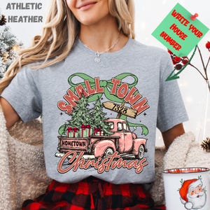 以下が含まれることがあります： クリスマス柄のグレーのアスレチックヘザーTシャツ。ピンクのトラックがクリスマスツリーとプレゼントを運んでいるグラフィックで、「Small Town Christmas」と「Hometown」の文字が表示されています。緑色の矢印の看板は7328番を指しています。