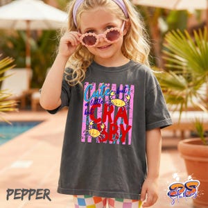 May include: Dark gray t-shirt with the text "Cute but a little CRA-BAY" in a colorful design featuring two yellow crabs. The shirt is worn by a young person wearing pink sunglasses and a headband.