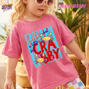 May include: A pink t-shirt with the text "Cute but a little Crabby" in yellow and red, featuring two yellow crab illustrations. The shirt has a crew neck and short sleeves.
