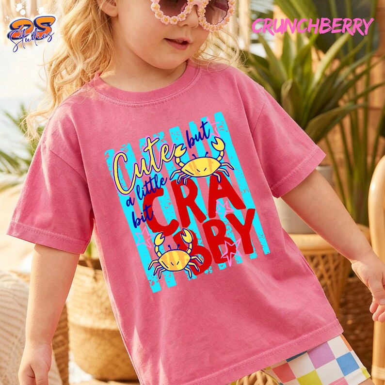 May include: A pink t-shirt with the text "Cute but a little Crabby" in yellow and red, featuring two yellow crab illustrations. The shirt has a crew neck and short sleeves.