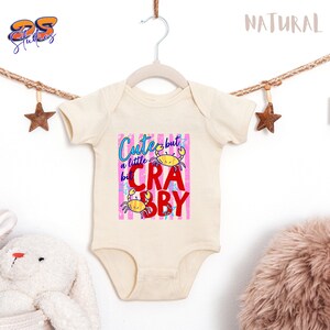 May include: Cream-colored baby onesie with a graphic that reads "Cute but a little Crabby." The design features two yellow crabs and text in blue and red against a pink striped background. The onesie is hanging on a white hanger.