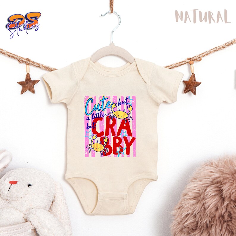 May include: Cream-colored baby onesie with a graphic that reads "Cute but a little Crabby." The design features two yellow crabs and text in blue and red against a pink striped background. The onesie is hanging on a white hanger.
