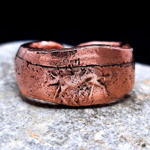 May include: A wide copper ring with a heavily textured, rugged surface. The metal features a reddish-brown metallic finish with deep, uneven indentations and a prominent horizontal fissure running across the center of the band's outer circumference.