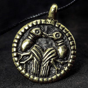 May include: A bronze-colored pendant with a circular design. The pendant features a detailed relief of two stylized bird-like figures facing each other, set within a textured border. The pendant hangs from a black cord.