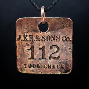May include: A square, copper-coloured pendant with the text "J.E.H. & SONS CO. 112 TOOL CHECK" hangs from a black cord. The pendant has a distressed, aged appearance, with a copper-coloured loop at the top for the cord.
