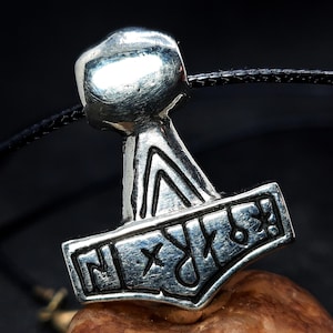 Puede incluir: Un colgante plateado con forma de martillo de Thor, Mjolnir, con símbolos rúnicos grabados. El colgante está suspendido de un cordón negro. El martillo descansa sobre una superficie marrón.