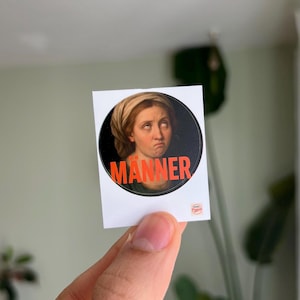 Könnte beinhalten: Ein weißer Aufkleber mit einem kreisförmigen Bild einer Frau mit missbilligendem Gesichtsausdruck auf schwarzem Hintergrund. Das Wort "MANNER" ist in fetten orangefarbenen Buchstaben am unteren Rand des Bildes gedruckt.