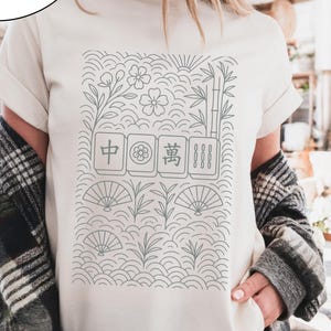 May include: White t-shirt with a gray graphic of Mahjong tiles, flowers, and fans. The text "COMFORT COLORS White" is printed on the top left. The shirt has rolled-up sleeves and is being worn.