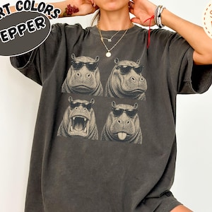 May include: A charcoal gray short-sleeve t-shirt featuring a graphic print of four hippos wearing black sunglasses. The hippos are arranged in a two-by-two grid, with one hippo shown with its mouth open and another sticking out its tongue.