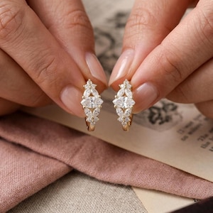 May include: A pair of gold hoop earrings with marquise-cut and round-cut white gemstones. The earrings are held up by a person's hands, showcasing the intricate design and sparkle of the jewelry. The earrings are a delicate and elegant accessory.