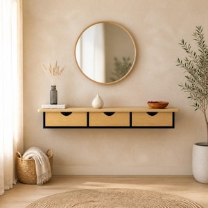 Peut inclure: Une console murale flottante dotée de trois tiroirs en bois clair avec des cadres noirs et des poignées découpées circulaires. Au-dessus de la console est suspendu un grand miroir rond avec un cadre fin en bois clair reflétant l'espace.