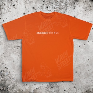Puede incluir: Camiseta naranja brillante con el texto blanco "channel ORANGE" centrado en el pecho. La camiseta tiene mangas cortas y cuello redondo. El fondo es una superficie gris texturizada.