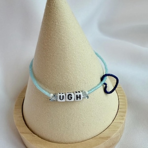 May include: A light blue beaded bracelet with the word "UGH" spelled out in white letter beads. The bracelet features a dark blue heart charm and clear crystal accents. The bracelet is displayed on a wooden stand.