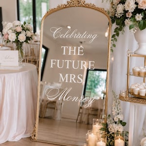 Könnte beinhalten: Ein hoher, gewölbter Standspiegel mit einem verzierten goldenen Rahmen. Die Glasoberfläche ist mit weißer Schrift versehen, die "Celebrating THE FUTURE MRS Monroy" lautet. Der Spiegel steht aufrecht und reflektiert den Veranstaltungsraum.