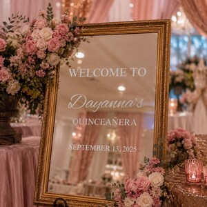 Puede incluir: Un espejo rectangular con un marco dorado ornamentado que presenta texto blanco que dice WELCOME TO Dayanna's QUINCEAÑERA SEPTEMBER 13, 2025. El espejo está decorado con arreglos de rosas rosas y blancas y follaje verde en las esquinas.