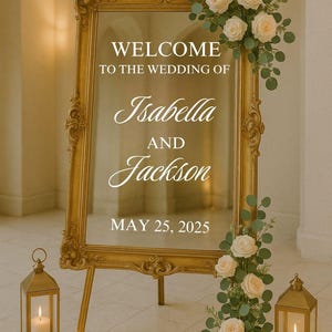 Puede incluir: Espejo dorado con el texto "WELCOME TO THE WEDDING OF Isabella AND Jackson MAY 25, 2025." Decorado con rosas blancas y vegetación. Dos faroles dorados con velas a cada lado.