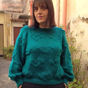 May include: A teal blue knitted jumper with a heart-shaped pattern. The jumper has a round neckline and long sleeves.