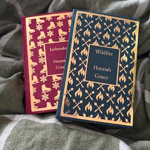 Peut inclure: Deux livres à couverture rigide avec des motifs dorés. L'un est bordeaux avec des patins à glace et des flocons de neige, intitulé "Icebreaker" par Hannah Grace. L'autre est bleu marine avec des flammes et des torches croisées, intitulé "Wildfire" par Hannah Grace.