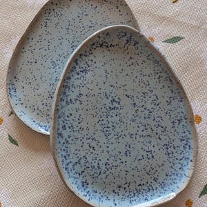 May include: Two light blue, oval ceramic plates with a speckled dark blue pattern. The plates have a slightly raised edge and a rustic, handmade appearance. The plates are placed on a cream tablecloth with a floral pattern.