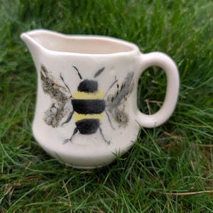 Könnte beinhalten: Eine kleine, cremefarbene Keramik-Sahnekännchen mit einem handgemalten Hummel-Design. Die Biene hat schwarze und gelbe Streifen mit grauen Flügeln. Das Sahnekännchen hat einen Griff und einen Ausguss. Das Sahnekännchen steht auf grünem Gras.