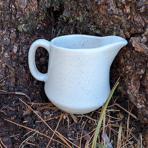 May include: A small, speckled white ceramic creamer with a curved handle and a pouring spout. The creamer is sitting on a bed of pine needles and dirt, with a textured tree trunk in the background. The creamer is a light grey colour.