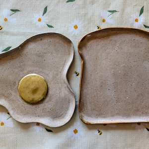 Peut inclure: Deux assiettes en céramique, une en forme d'œuf au plat avec un jaune d'œuf jaune, l'autre en forme de tranche de pain grillé. Les deux assiettes sont mouchetées et de couleurs neutres, parfaites pour servir le petit-déjeuner ou des collations.