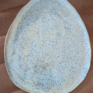 May include: A speckled ceramic serving platter with an organic shape. The platter has a light blue base with dark blue speckles and a slightly raised edge. The design is simple and modern, suitable for serving appetizers or small dishes.
