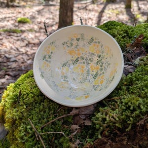 Peut inclure: Un bol en céramique crème avec un motif floral jaune et vert. Le bol est rond avec un bord légèrement évasé. Le motif floral recouvre l'intérieur. Le bol est posé sur un lit de mousse verte.