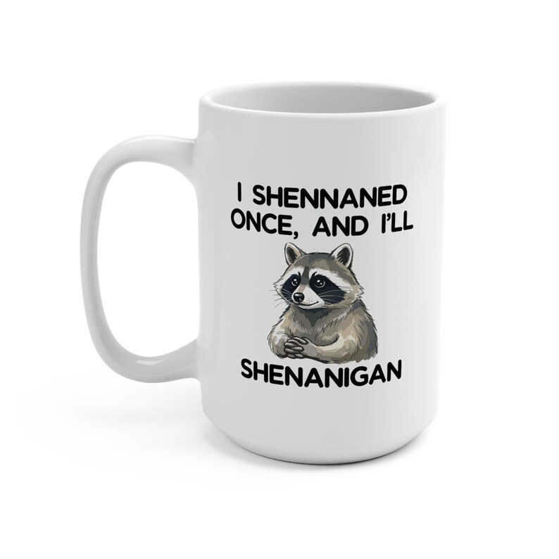 May include: White ceramic mug with a black handle. It features a raccoon illustration and the text "I Shennaned Once, And I'll Shenanigan". The raccoon is depicted with folded paws.