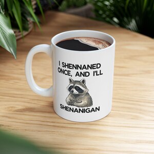May include: White ceramic coffee mug with a black interior filled with coffee. The mug features a raccoon illustration and the text "I Shennaned Once, And I'll Shenanigan."