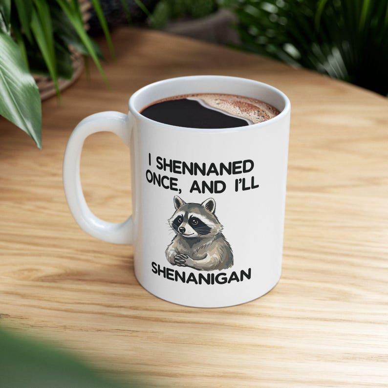 May include: White ceramic coffee mug with a black interior filled with coffee. The mug features a raccoon illustration and the text "I Shennaned Once, And I'll Shenanigan."