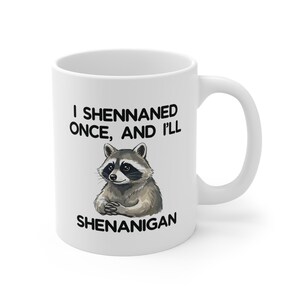 May include: White ceramic mug with a black handle. The mug has a raccoon illustration and the text "I Shennaned Once, And I'll Shenanigan". The raccoon is depicted with gray and brown fur.