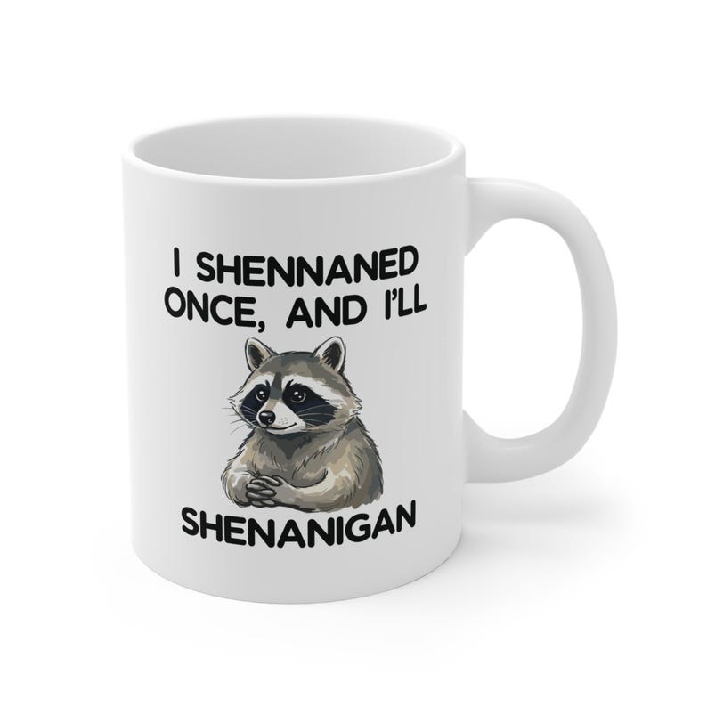 May include: White ceramic mug with a black handle. The mug has a raccoon illustration and the text "I Shennaned Once, And I'll Shenanigan". The raccoon is depicted with gray and brown fur.