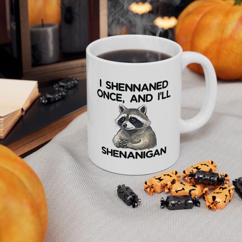 May include: White ceramic mug featuring a raccoon illustration and the text "I Shennaned Once, And I'll Shenanigan". The mug contains a dark liquid. Halloween decorations are visible in the background.