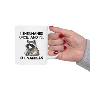 May include: White ceramic mug featuring a raccoon illustration and the text "I Shennaned Once, And I'll Shenanigan". The raccoon is grey and white with folded arms. The mug has a white handle.