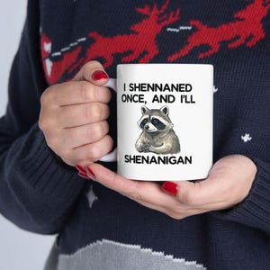May include: White ceramic mug with a raccoon illustration and the text "I Shennaned Once, And I'll Shenanigan." The raccoon is shown with folded arms. The mug is held by a person wearing a dark blue sweater.