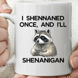 May include: White ceramic mug with a raccoon illustration and the text "I Shennaned Once, And I'll Shenanigan" in black. The raccoon has a black mask and is in a seated position.