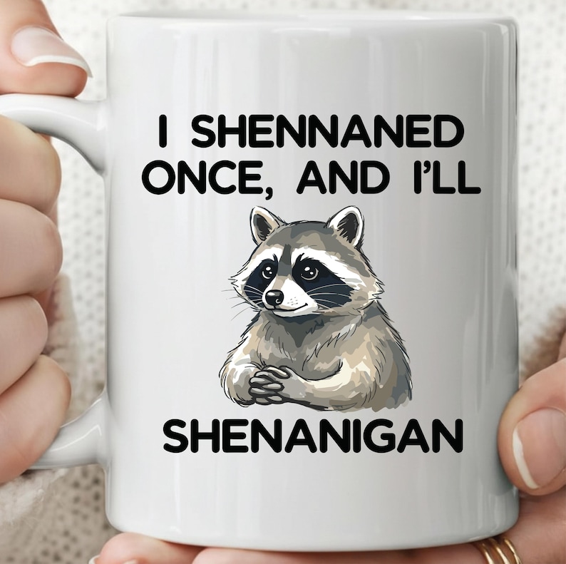 May include: White ceramic mug with a raccoon illustration and the text "I Shennaned Once, And I'll Shenanigan" in black. The raccoon has a black mask and is in a seated position.