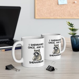 May include: Two white ceramic mugs with the text "I Shennaned Once, And I'll Shenanigan" and a raccoon illustration. The mugs are on a light-coloured surface with a laptop and office supplies in the background.