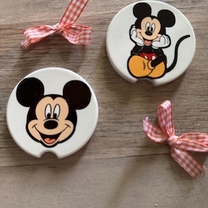 May include: Two white ceramic coasters with a cartoon image of Mickey Mouse. One coaster features Mickey Mouse sitting with his hands on his chin, the other coaster features Mickey Mouse's face. Both coasters have a red and white checkered ribbon tied around them.