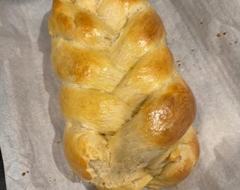 May include: A braided loaf of bread, golden brown and slightly glossy, sits on a white parchment-lined baking sheet.