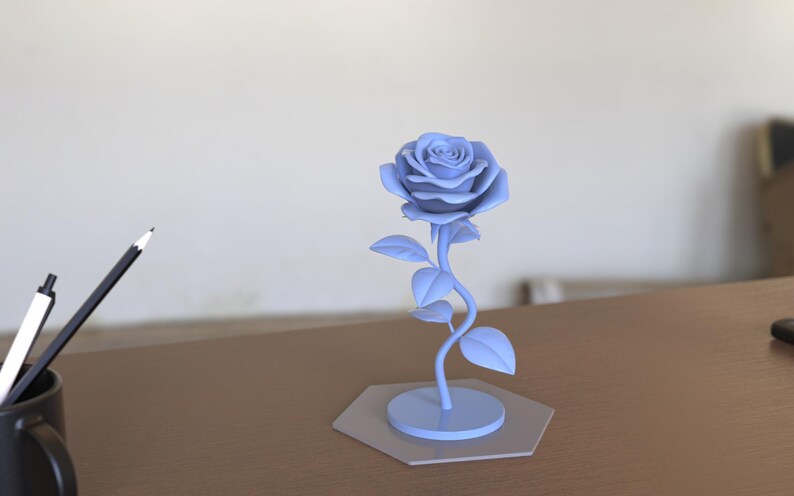 May include: A light blue, single-stem rose sculpture featuring a detailed, multi-petaled flower head, a curved stem with several leaves, and a circular base attached to a flat, light gray hexagonal platform, presented as a solid decorative object.