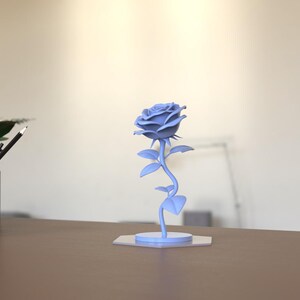 May include: A light blue rose sculpture featuring a detailed flower head with layered petals, a slender stem with several leaves, and a circular base. The object is rendered in a solid matte light blue color and stands upright on a flat surface.
