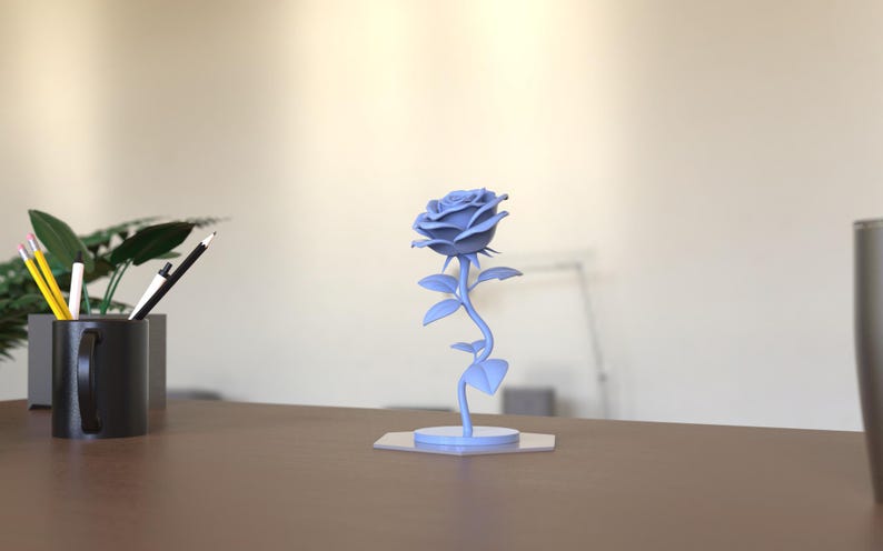 May include: A light blue rose sculpture featuring a detailed flower head with layered petals, a slender stem with several leaves, and a circular base. The object is rendered in a solid matte light blue color and stands upright on a flat surface.