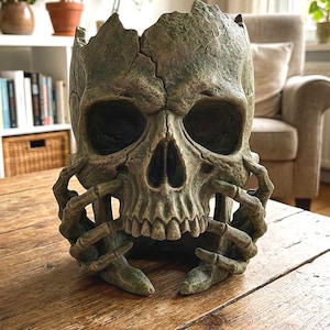 Peut inclure: Un pot de fleurs en forme de crâne, de couleur verte et vieillie, avec des fissures visibles. Des mains squelettiques soutiennent le crâne, créant une esthétique gothique unique. Idéal pour Halloween ou pour ajouter une touche effrayante à la décoration.
