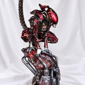 May include: A metallic red alien creature figurine in a crouching position, featuring a long segmented tail, exposed mechanical details, and glowing green accents. The creature is perched atop a textured, dark, industrial base with pipes.