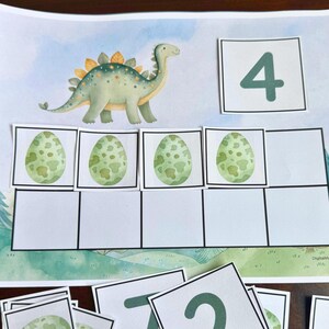 May include: A colorful educational print featuring a watercolor illustration of a green dinosaur and four green eggs. The number 4 is displayed in a square, with additional number cards and egg cards scattered below.